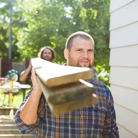 Two carpenters carrying lumber past a fence on a residential job
